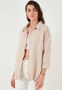 Chemise boutonnée en beige clair, fabriquée en tissu texturé. Caractéristiques : un col, des manches longues retroussées, et une coupe décontractée avec un ourlet droit.