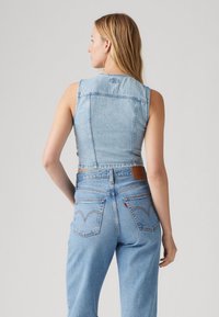 Světlomodrá krátká džínová vesta s přiléhavým střihem. Má rovný zadní díl, viditelné švy a hnědý kožený záplatu v pase.