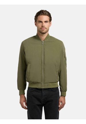 Mężczyzna o krótkich brązowych włosach, ubrany w oliwkowo-zieloną kurtkę bomberkę zapinaną na zamek, stojący na tle jednolitego jasnego tła.