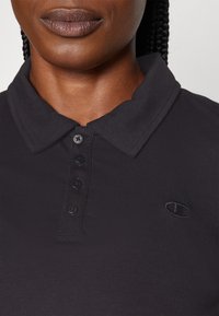 Närbild av en person som bär en svart polo med en fyraknappslåsning och en liten svart broderad Champion-logga på bröstet.