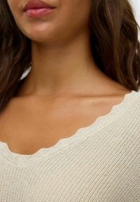 Top beige chiaro con scollatura a scallop, tessuto testurizzato e un leggero luccichio. Ingrandimento della zona del petto, che mostra il tono della pelle e i dettagli del tessuto.