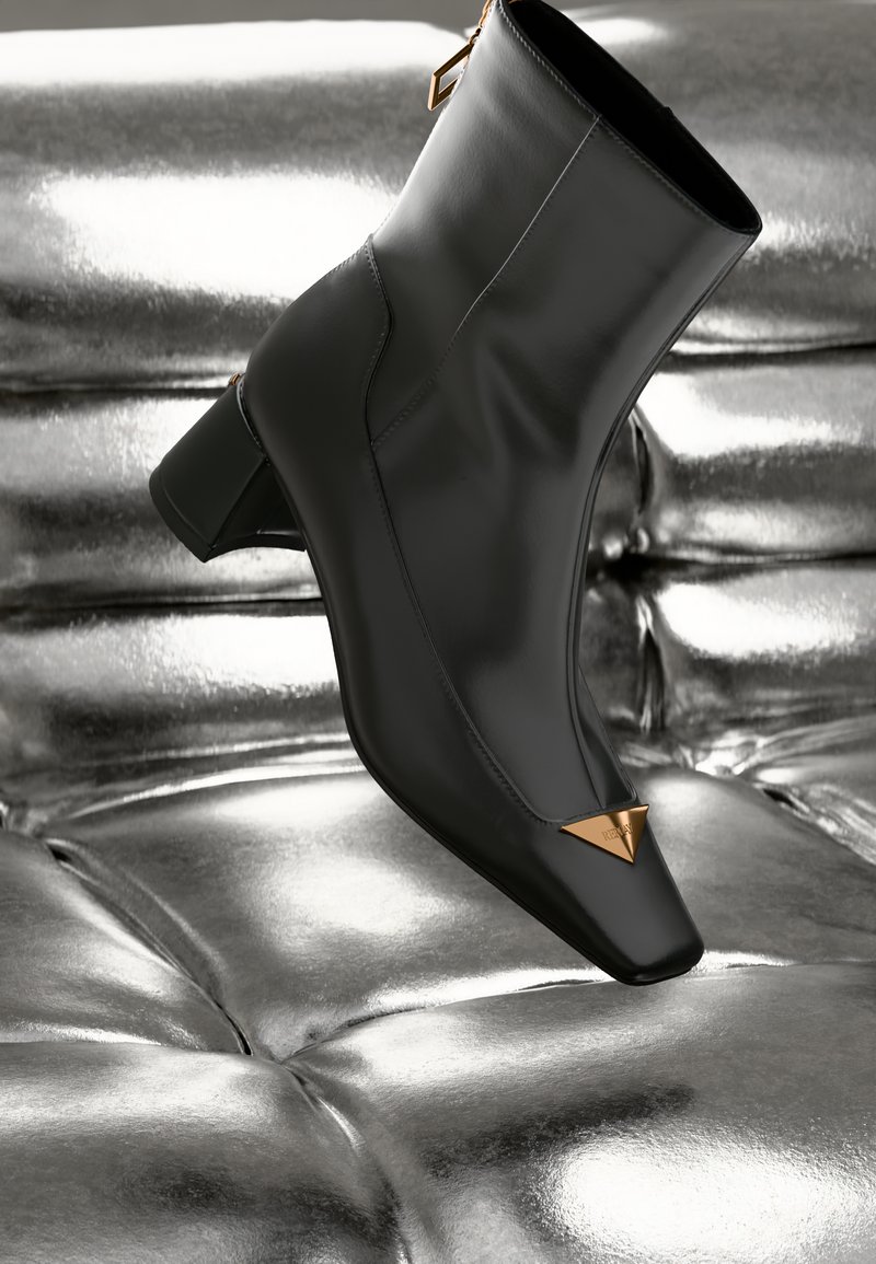 Stivaletto in pelle nera con punta appuntita, accento triangolare dorato sul davanti e tacco a blocco, su uno sfondo metallico argento.