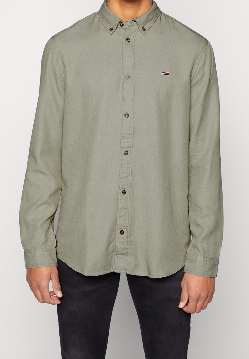 Homme portant une chemise boutonnée manches longues vert olive clair avec un petit logo rouge, blanc et bleu sur la poitrine, associée à un pantalon noir.