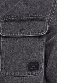 Dunkelgraues Jeanshemd mit Klappentasche und schwarzem Druckknopf, versehen mit einem kleinen schwarzen Stofflabel mit weißem Text unten rechts.