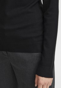 Maglione nero in maglia con design aderente, polsini a coste e una texture liscia. Abbinato a pantaloni in tessuto grigio scuro, che mettono in risalto una silhouette elegante.