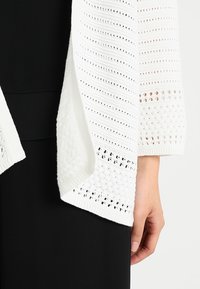Vit stickad cardigan med ett texturerat mönster och öppen front. Har partiella nätdetaljer och vida ärmar, lagd över svart klädsel.