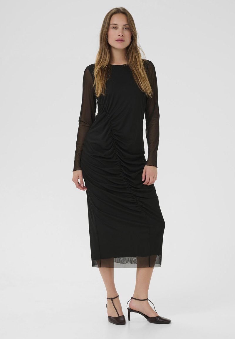 Black mid-length dress with long sheer sleeves, ruched detailing on the left side, smooth texture, and an elegant silhouette. Paired with black heels.