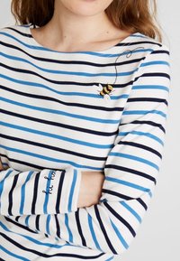 Maglia a maniche lunghe a righe blu e navy su tessuto bianco. Presenta un design ricamato di un'ape e un polsino con il testo "be happy" in blu.