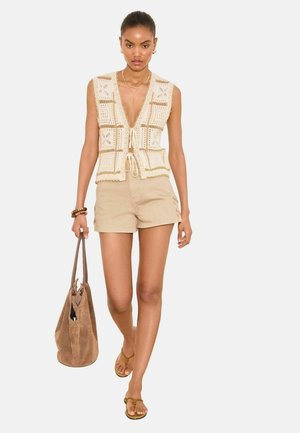 Žena kráča, nosiac béžovú háčkovanú vestu, hnedé šortky, hnedé sandále a nesie veľkú semišovú kabelku.