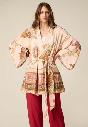 Donna che indossa una giacca in stile kimono floreale con maniche ampie e cintura, abbinata a pantaloni larghi rossi uniti, in piedi con le mani sui fianchi.