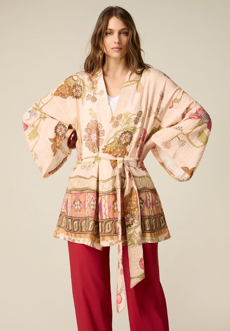 Woman wearing a floral kimono-style jacket with wide sleeves and a belt, paired with solid red wide-leg pants, standing with hands on hips.