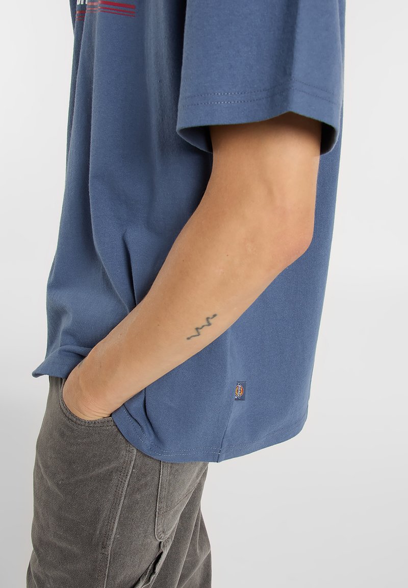 Blue cotton T-shirt with short sleeves, featuring a small logo patch at the hem. Worn with dark gray pants, hands in pockets.