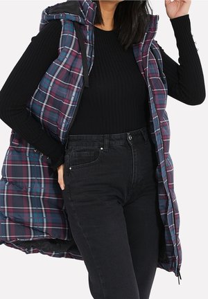 Femme portant un pull noir côtelé, un jean noir taille haute et une doudoune sans manches à carreaux violet-bleu avec capuche.
