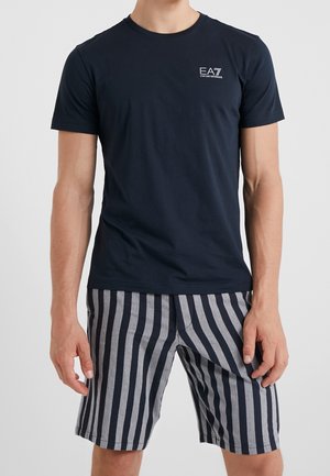 Mann trägt ein marineblaues EA7 Emporio Armani T-Shirt und schwarz-grau gestreifte Shorts mit vertikalen Streifen, steht vor einem schlichten, hellen Hintergrund.