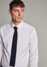 Camisa blanca de vestir con un collar estructurado, combinada con una corbata texturada en color azul marino. Presenta mangas largas y una silueta ajustada.