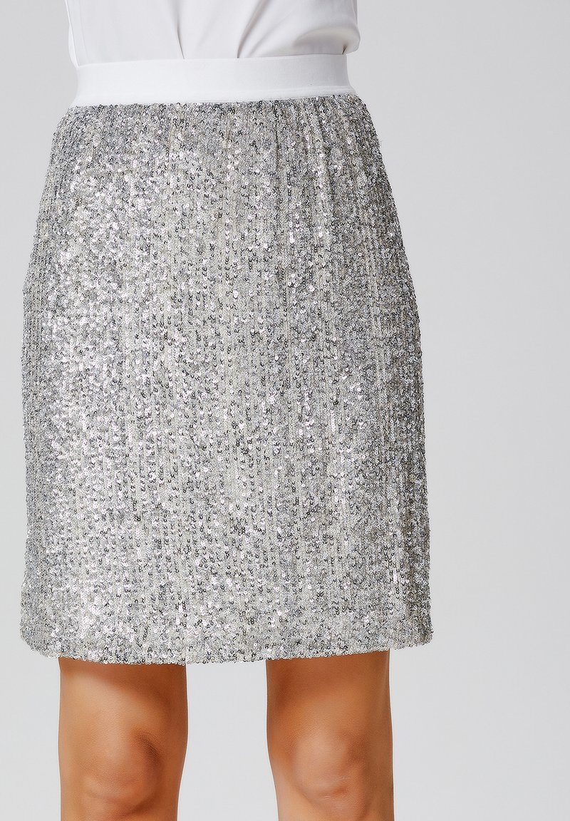 Jupe en sequins argentés avec une ceinture élastique blanche. Le tissu a une finition texturée et scintillante, et la jupe arrive au-dessus des genoux.