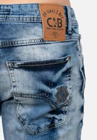 Jeans en denim de couleur bleu clair avec une texture déchirée. Présente une pièce en cuir brun avec logo, des coutures contrastées et une poche arrière décorative.
