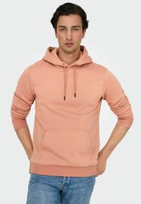 Ljusrosa hoodie i mjukt tyg, med en framficka, dragsko i huvan och ribbade mudd och avslut. Bärs med ljusblå jeans.