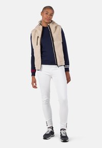Beige Fleece-Weste mit Kapuze, marineblauer Pullover, weiße Skinny-Jeans und schwarze Sneakers; hat Reißverschlüsse und gestreifte Bündchen.