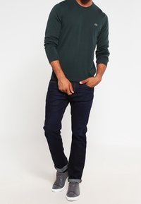 Mann trägt einen dunkelgrünen Langarmpullover, dunkelblaue Jeans und graue High-Top-Sneaker und steht mit einer Hand in der Tasche.