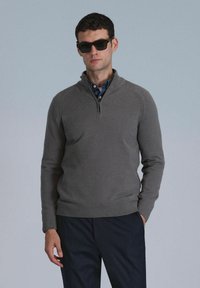 Grijze pullover trui met een half-rits kraag, gemaakt van gebreid materiaal. Het model draagt een donkere zonnebril en een blauw geruit shirt eronder.