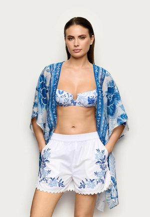 Donna con top bikini floreale blu e bianco, copricostume stile kimono abbinato e pantaloncini bianchi con ricamo floreale blu. Mani nelle tasche.