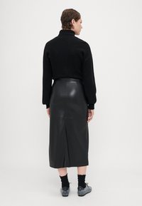 Suéter preto de malha com gola alta, combinado com uma saia midi de couro preta com uma fenda nas costas, usando calçado cinza e meias pretas.