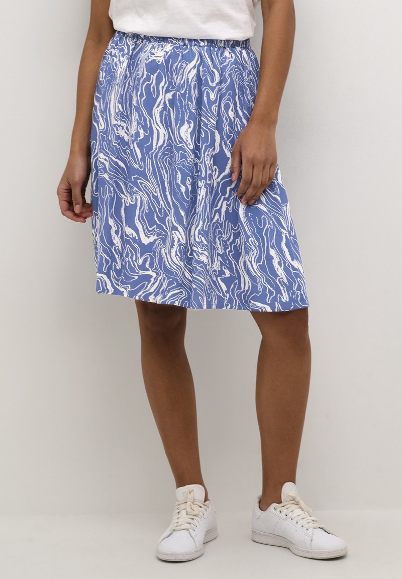 Femme portant une jupe mi-longue aux genoux avec un motif abstrait bleu et blanc, un haut blanc et des baskets blanches, debout devant un fond uni.