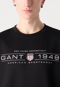 Svart hettegenser med rund hals, med hvit tekst og et emblem som viser røde og blå aksenter, merket "GANT 1949" og "AMERICAN SPORTSWEAR."