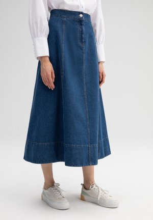 Frau trägt einen blauen Denim A-Linien Midirock, weißes Hemd mit Knöpfen und weiße Sneaker mit hellgrünen Details.
