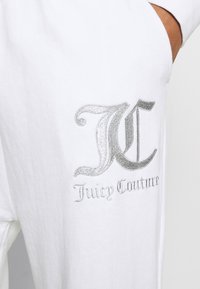 Witte katoenen kledingstuk met een zilveren geborduurd logo met een gestileerde "JC" en de tekst "Juicy Couture" eronder.