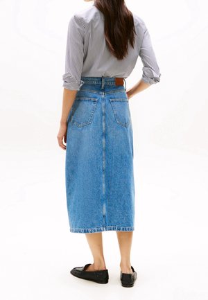 Femme aux cheveux longs portant une chemise rayée, une jupe en jean bleue mi-longue et des chaussures plates noires, debout avec une main sur la hanche.