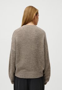 Žena s rovnými blond vlasy, oblečená v volném hnědém pleteném svetru a černých kalhotách, zobrazená zezadu na jednobarevném pozadí.