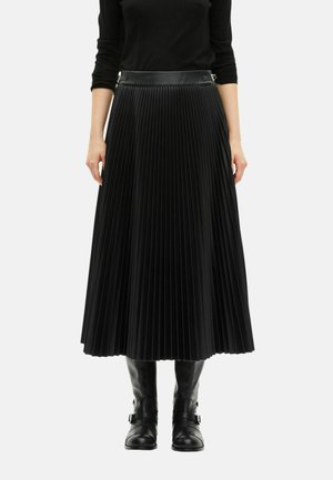 Γυναίκα με μαύρη πλισέ midi φούστα με λεπτομέρειες από πόρπες στη μέση και μαύρες δερμάτινες μπότες μέχρι το γόνατο σε άσπρο φόντο.