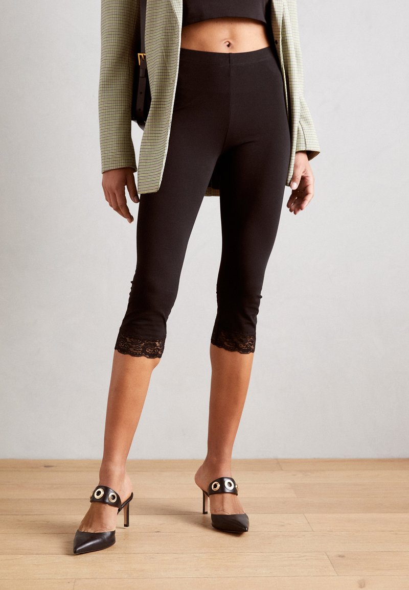 Sorte leggings med spidse kanter i kanten, parret med stilfulde sorte hæle med et rundt designelement og en stiletto hæl.