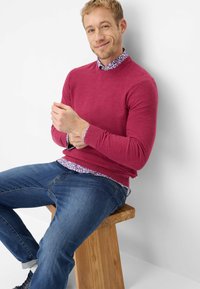 Bordeauxroter Pullover mit Rundhalsausschnitt über einem Hemd mit floralem Muster, kombiniert mit blauen Jeans. Sitzgelegenheit: Holzschemel.