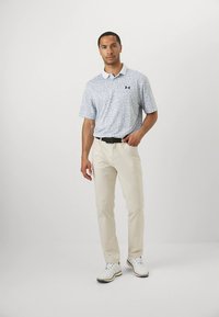 Muž oblečený v svetlošedom polu s vzorom, béžových nohaviciach, čiernom opasku a bielych golfových topánkach, stojaci na bielom pozadí.