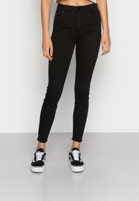 Jeans ajustados negros de mezclilla, con una cintura alta, diseño estándar de cinco bolsillos y cierre de botón, combinados con zapatillas negras.