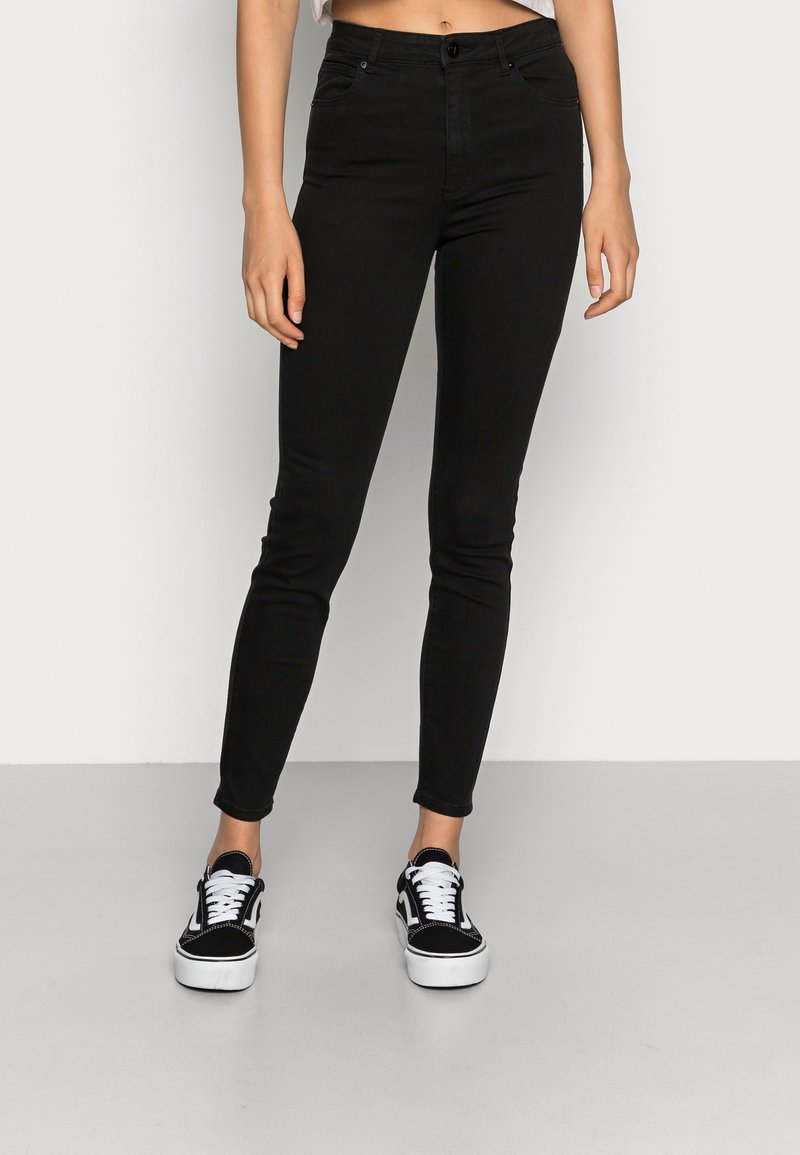 Jeans ajustados negros de mezclilla, con una cintura alta, diseño estándar de cinco bolsillos y cierre de botón, combinados con zapatillas negras.