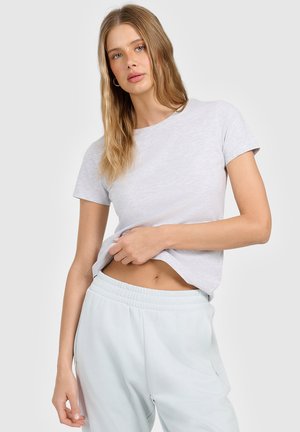 Młoda kobieta o długich blond włosach, ubrana w jasnoszarą koszulkę z krótkim rękawem i jasne spodnie dresowe, lekko podnosząca koszulkę.