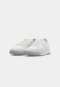 Paire de baskets basses blanches avec un design à lacets et un marquage discret sur le côté, affichées sur un fond gris clair uni.