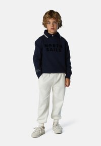 Felpa blu navy con logo "NORTH SAILS", colletto e polsini bianchi, abbinata a pantaloni casual bianchi e sneaker chiare.