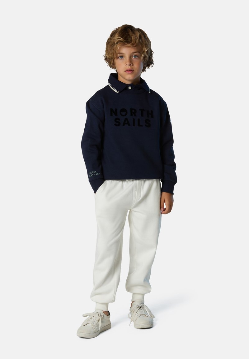 Felpa blu navy con logo "NORTH SAILS", colletto e polsini bianchi, abbinata a pantaloni casual bianchi e sneaker chiare.