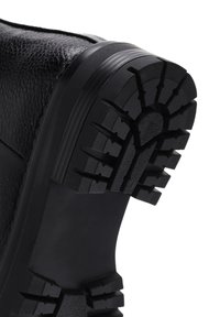 Suela de goma negra con un patrón de tracción distintivo, parte superior de cuero texturizado y talón reforzado. Presenta un acabado mate y suave.