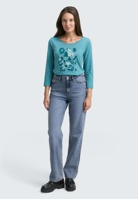 Türkises Langarmshirt mit grafischem Druck, kombiniert mit hochgeschnittenen hellblauen Jeans und schwarzen klobigen Plateauschuhen.