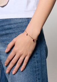 Złota bransoletka łańcuszkowa z małym czerwonym serduszkiem i cylindrycznym akcentem, noszona na dłoni na tle niebieskiego denimu.