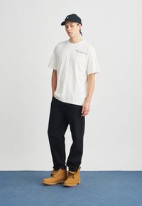 Biała bawełniana koszulka z nadrukiem, czarne spodnie i żółto-brązowe buty. Czarna czapka dopełnia casualowy strój na jednolitym tle.