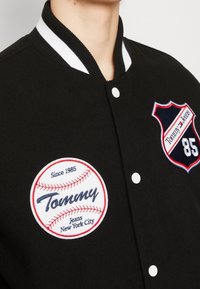 Czarna kurtka varsity z białym prążkowanym kołnierzem, zdobiona czerwonym i białym wyszywanym patch'em baseballowym oraz logo tarczy z detalem "85".