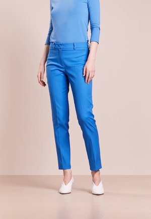 Persona vistiendo pantalones ajustados de color azul brillante, camiseta de manga larga a juego en azul claro, y zapatos blancos de punta afilada, de pie sobre un suelo beige.