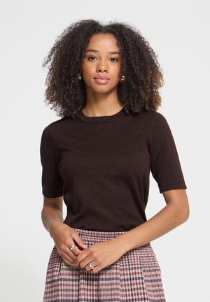 Femme aux cheveux bouclés portant un haut marron foncé à manches courtes et une jupe à carreaux, debout les mains jointes à la taille sur fond blanc.